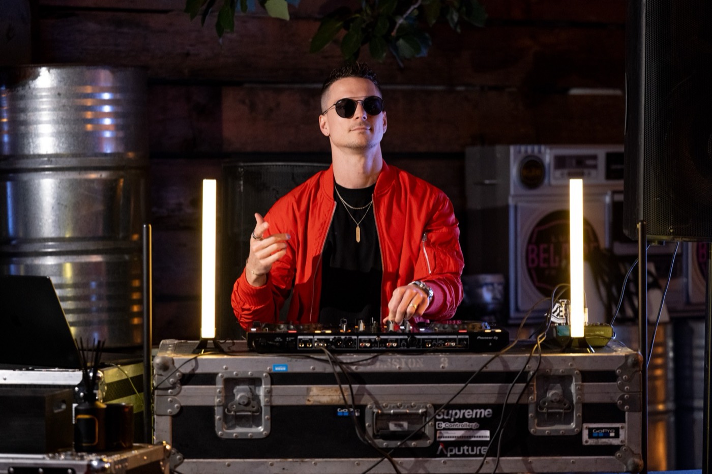 DJ in a red jacket performing behind illuminated light bars in a dark venue