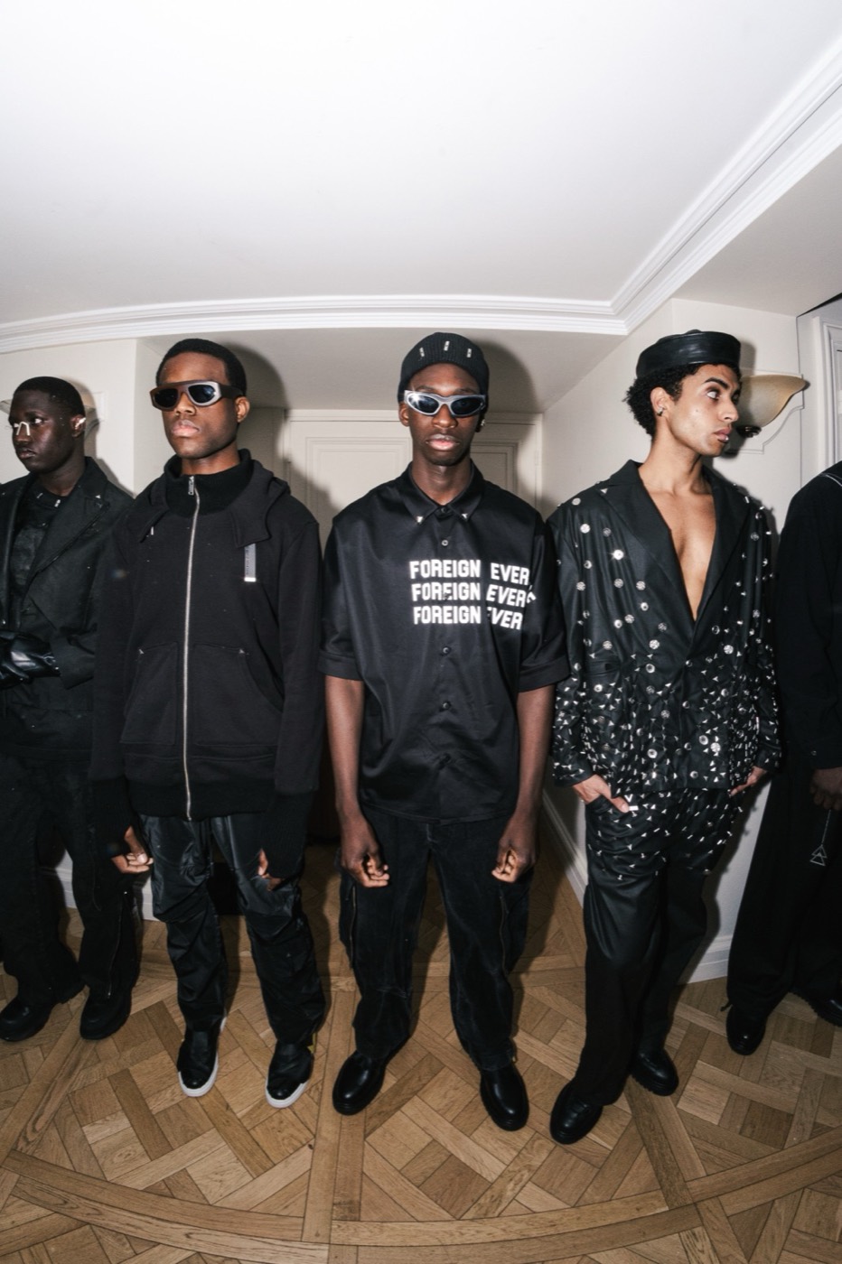 Five men in black outfits posing in a row at a fashion event